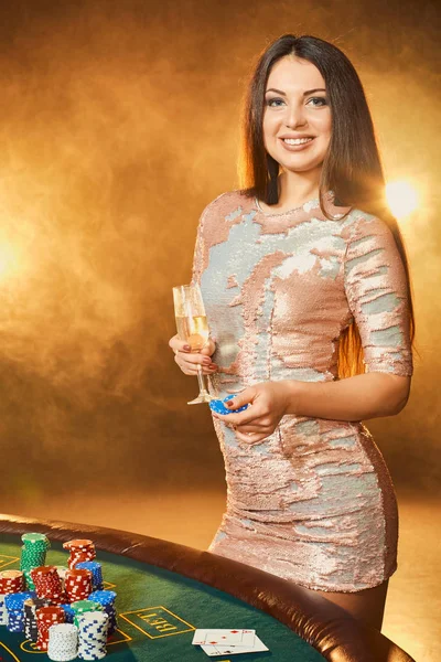 Preciosa joven en vestido de noche de pie cerca de la mesa de póquer con copa de champán — Foto de Stock