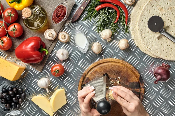 Las manos femeninas frotaron queso rallado en la pizza, ingredientes para cocinar pizza en la mesa de metal, vista superior — Foto de Stock