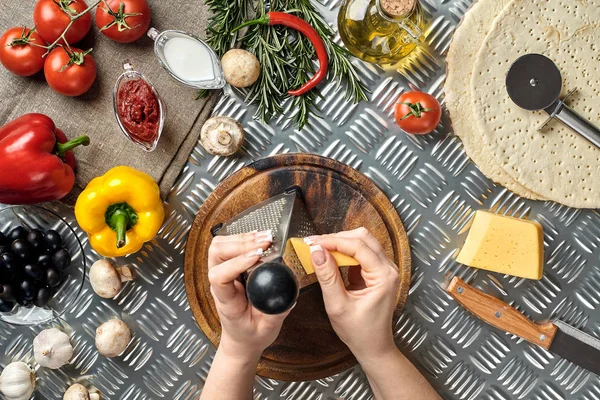 Las manos femeninas frotaron queso rallado en la pizza, ingredientes para cocinar pizza en la mesa de metal, vista superior — Foto de Stock