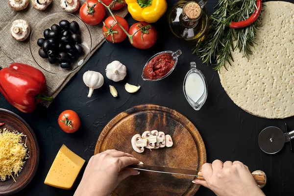 Las setas femeninas cortadas a mano en el tablero de madera en la mesa de la cocina, alrededor de los ingredientes de mentira para la pizza: verduras, queso y especias . — Foto de Stock