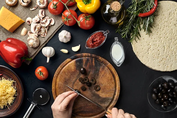 Las aceitunas femeninas cortadas a mano sobre tabla de madera en la mesa de la cocina, alrededor de la mentira ingredientes para la pizza: verduras, queso y especias . — Foto de Stock