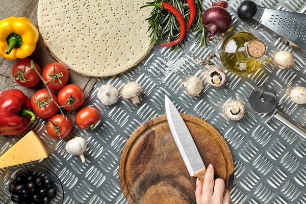 Mano femenina con cuchillo en la mesa de la cocina de metal, alrededor de los ingredientes de mentira, verduras y especias para pizza . — Foto de Stock