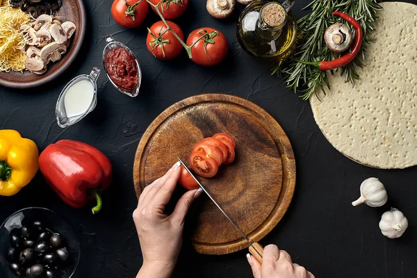 Hembra tomates cortados a mano en tablero de madera en la mesa de la cocina, alrededor de los ingredientes de mentira para pizza: verduras, queso y especias . — Foto de Stock