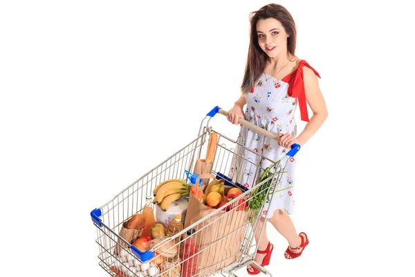 Vista de ángulo alto de la niña sonriendo a la cámara mientras empuja un carrito de compras lleno de comestibles aislados sobre fondo blanco . — Foto de Stock