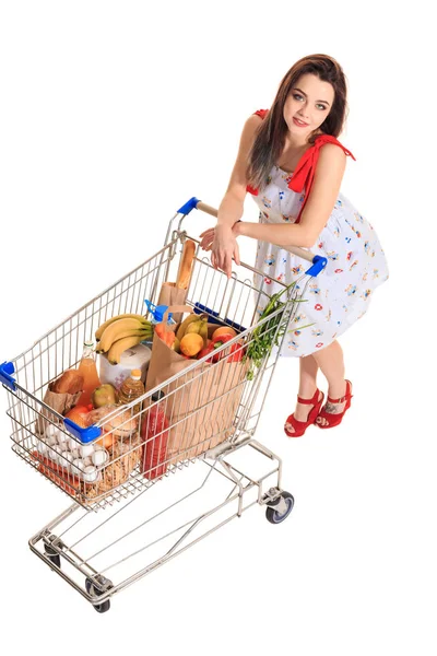 Wysoki kąt widzenia dziewczyna uśmiechając się aparatu podczas pchania zakupy koszyka pełnego spożywcze na białym tle. — Zdjęcie stockowe