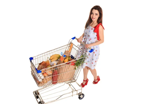 Vista de ángulo alto de la niña sonriendo a la cámara mientras empuja un carrito de compras lleno de comestibles aislados sobre fondo blanco . — Foto de Stock
