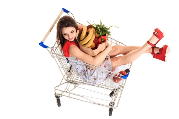 Vista de ángulo alto de la niña sonriendo a la cámara mientras sostiene bolsas de comestibles y sentado en el carro de la compra aislado en blanco — Foto de Stock