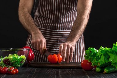 Adam ahşap bir masa salataya hazırlanıyor. Mens eller siyah arka plan üzerine bir salata domates kesmek