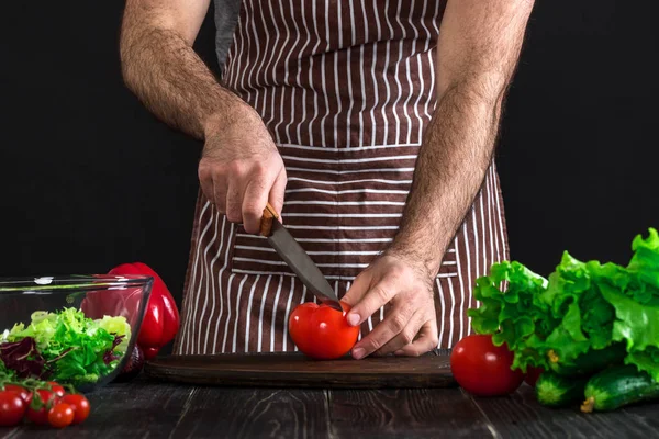 Adam ahşap bir masa salataya hazırlanıyor. Mens eller siyah arka plan üzerine bir salata domates kesmek