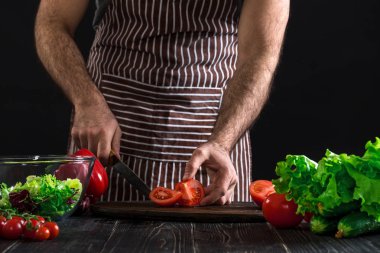 Adam ahşap bir masa salataya hazırlanıyor. Mens eller siyah arka plan üzerine bir salata domates kesmek