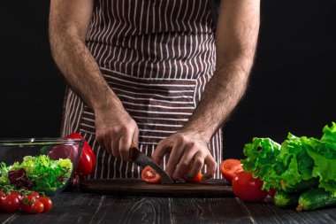Adam ahşap bir masa salataya hazırlanıyor. Mens eller siyah arka plan üzerine bir salata domates kesmek