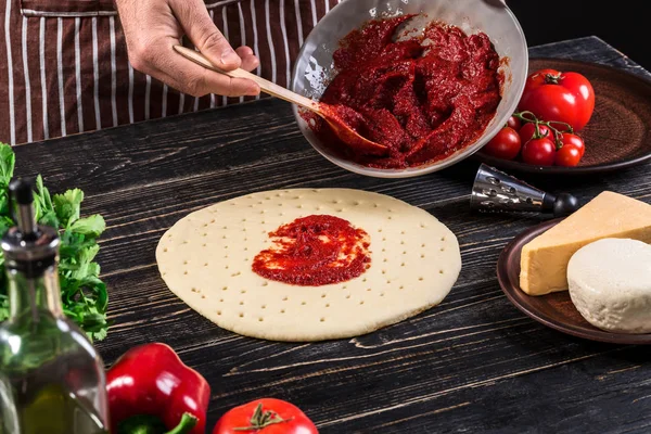 Μια αρσενική χέρι εξαπλώνεται ντομάτα πουρέ στη μια βάση της πίτσας με κουτάλι σε ένα παλιό ξύλινο φόντο — Φωτογραφία Αρχείου