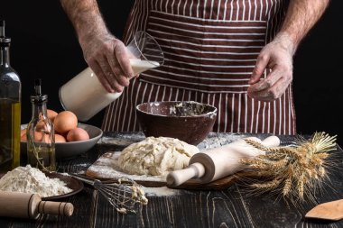 Baker plastik şişe süt kasenin içine dökme üniformalı. Bir avuç un yumurta rustik bir mutfak ile.