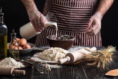 Baker plastik şişe süt kasenin içine dökme üniformalı. Bir avuç un yumurta rustik bir mutfak ile.