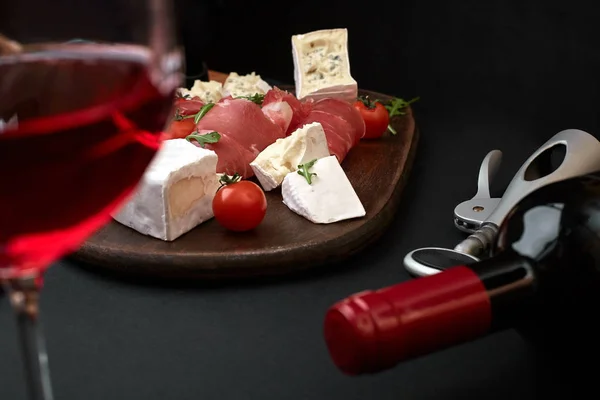 Delicious appetizer to wine - ham, cheese, baguette slices, tomatoes, served on a wooden board, and glass with red wine on black surface