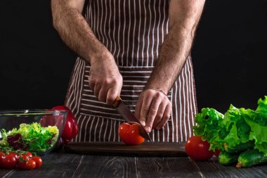 Adam ahşap bir masa salataya hazırlanıyor. Mens eller siyah arka plan üzerine bir salata domates kesmek