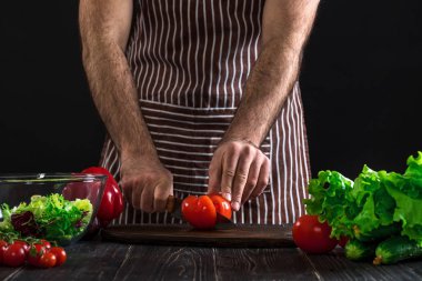 Adam ahşap bir masa salataya hazırlanıyor. Mens eller siyah arka plan üzerine bir salata domates kesmek