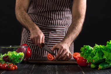 Adam ahşap bir masa salataya hazırlanıyor. Mens eller siyah arka plan üzerine bir salata domates kesmek