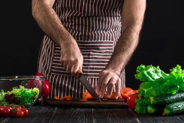 Genç ev aşçı adam havuç mutfak bıçağı ile dilimleme önlük. Mens eller havuç siyah arka plan üzerine bir salata yapmak için kesti. Sağlıklı gıda kavramı
