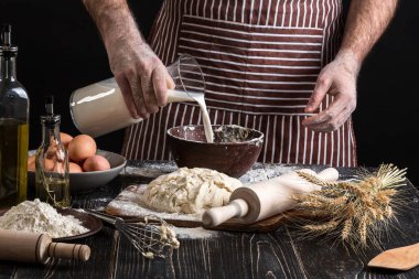 Baker plastik şişe süt kasenin içine dökme üniformalı. Bir avuç un yumurta rustik bir mutfak ile.