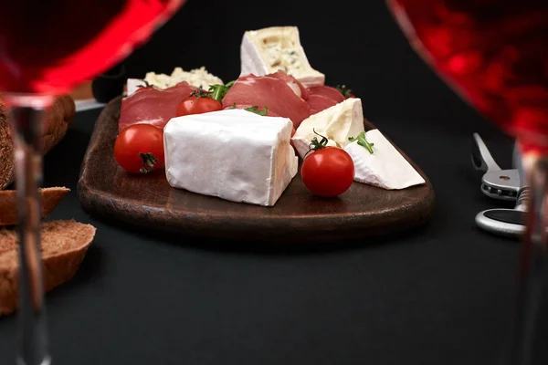 Prosciutto, salami, baguette slices, tomatoes and nutson rustic wooden board, two glasses of red wine over black background — Stock Photo, Image