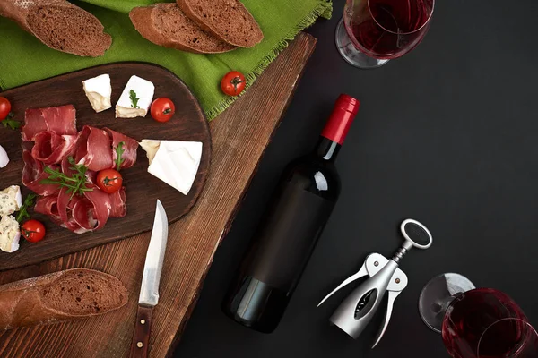 Red wine, cheese, cherry tomato, bread and prosciutto on wooden board over black backdrop, top view, copy space