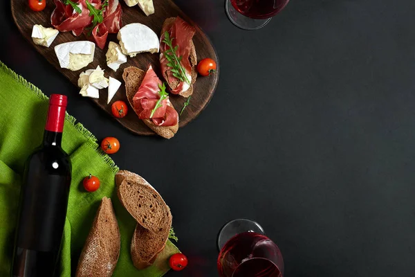 Delicious appetizer to wine - ham, cheese, baguette slices, tomatoes, served on a wooden board, and glass with red wine on black surface