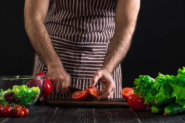 Adam ahşap bir masa salataya hazırlanıyor. Mens eller siyah arka plan üzerine bir salata domates kesmek