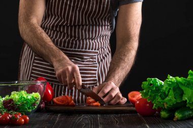 Adam ahşap bir masa salataya hazırlanıyor. Mens eller siyah arka plan üzerine bir salata domates kesmek