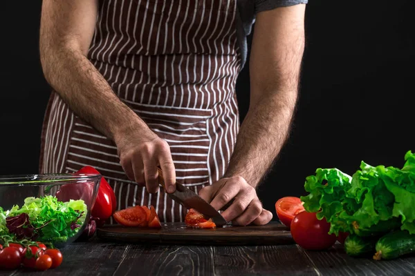 Hombre preparando ensalada en una mesa de madera. Manos para hombre cortar el tomate para hacer una ensalada sobre fondo negro — Foto de Stock