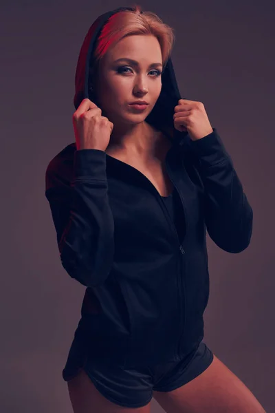 Giovane bella donna indossa un cappuccio nero e pantaloncini su sfondo scuro — Foto Stock