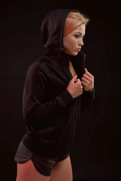 Beautiful seductive sporty girl with the slim sexy figure in the black hoodie, sport underwear and little black shorts is posing in the studio — Stock Photo, Image