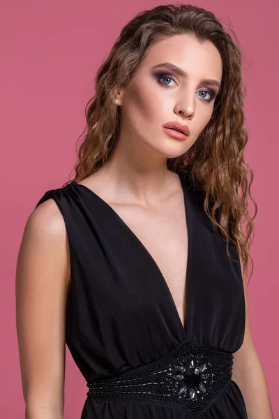 Hermosa chica de moda con el pelo largo y rizado en un vestido negro en el estudio sobre un fondo rosa . —  Fotos de Stock