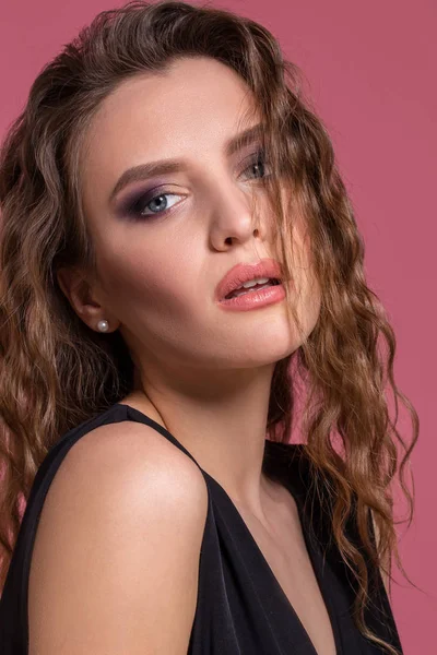 Hermosa chica de moda con el pelo largo y rizado en un vestido negro en el estudio sobre un fondo rosa . — Foto de Stock