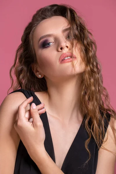 Schöne modische Mädchen mit langen lockigen Haaren in einem schwarzen Kleid im Studio auf rosa Hintergrund. — Stockfoto