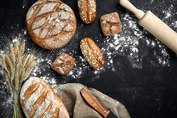 Hausgemachtes Roggenbrot, bestreut mit Mehl und verschiedenen Körnern und Samen auf schwarzem Hintergrund mit Ähren aus Weizen oder Roggen und Hafer. — Stockfoto