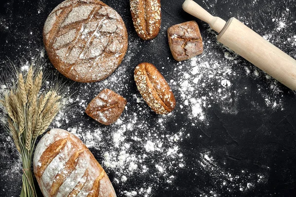 Hausgemachtes Roggenbrot, bestreut mit Mehl und verschiedenen Körnern und Samen auf schwarzem Hintergrund mit Ähren aus Weizen oder Roggen und Hafer. — Stockfoto