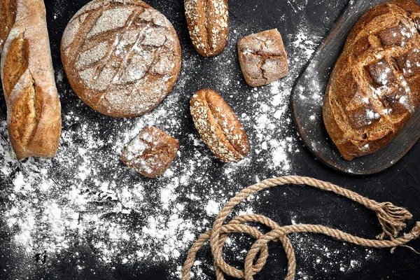 Pâine de secară de casă presărată cu făină și diverse cereale și semințe pe un fundal negru cu spikelets de grâu sau secară și ovăz . — Fotografie, imagine de stoc