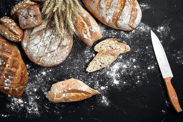 Pane di segale fatto in casa cosparso di farina e vari cereali e semi su fondo nero con spighe di frumento o segale e avena . — Foto Stock