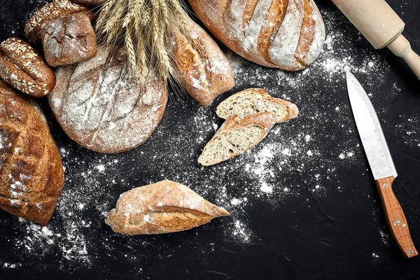 Pane di segale fatto in casa cosparso di farina e vari cereali e semi su fondo nero con spighe di frumento o segale e avena . — Foto Stock