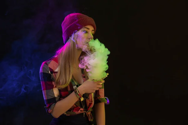 Выпендривающаяся девушка. Молодая хипстерша испаряет электронную сигарету на черном фоне. Хип-хоп стиль. Снимок студии — стоковое фото