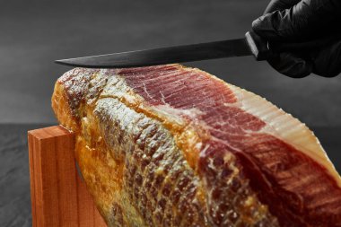 Siyah eldivenli bir aşçının eli jamoner standında domuz budu bıçağıyla bir parça jamon kesiyor. Yakın plan çekim. Yan görünüm.