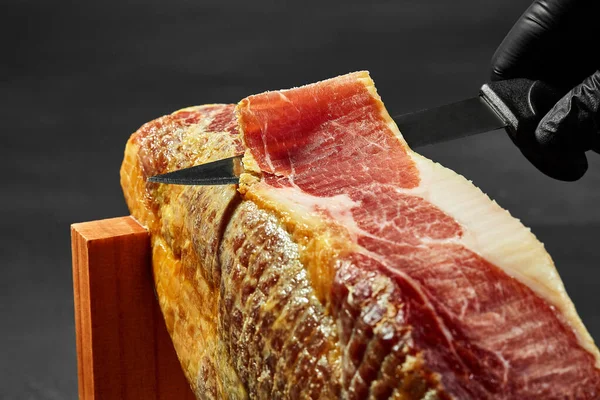 La mano de un cocinero en un guante negro está cortando una pieza de jamón con un cuchillo de una pierna de cerdo en un puesto de jamón. Cerrado el tiro. Vista lateral. — Foto de Stock