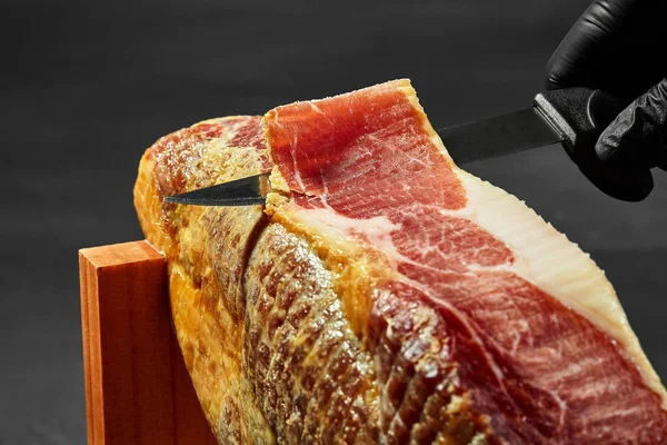Hand of a cook in a black glove is cutting out a piece of jamon with a knife from a pork leg on a jamonera stand. Close-up shot. Side view. — Stock Image Hand of a cook in a black glove is cutting out a piece of jamon with a knife from a pork leg on a jamonera stand. Close-up shot. Side view. Stock Photo