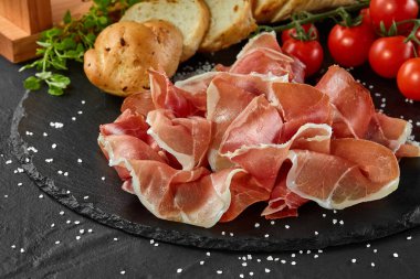 Jamon, jamoneranın üstünde domuz bacağından oyulmuş, kiraz domatesleri, baget, otlar, siyah tahtaya baharatlar. Karanlık arka plan. Yakın plan. Yan görünüm.
