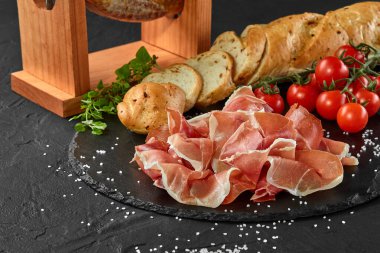 Jamon, jamoneranın üstünde domuz bacağından oyulmuş, kiraz domatesleri, baget, otlar, siyah tahtaya baharatlar. Karanlık arka plan. Yakın plan. Yan görünüm.