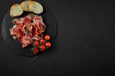 Dilimlenmiş jamon, vişneli domates, baget, siyah taşlı tahta üzerinde koyu gri bir arka plan. Yakın plan çekim. Üst Manzara. Boşluğu kopyala.