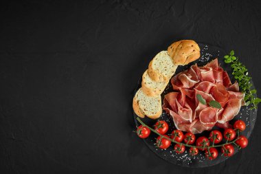 Dilimlenmiş jamon, kiraz domatesleri, baget, otlar ve baharatlar siyah gri arka planda. Yakın plan çekim. Üst görünüm.