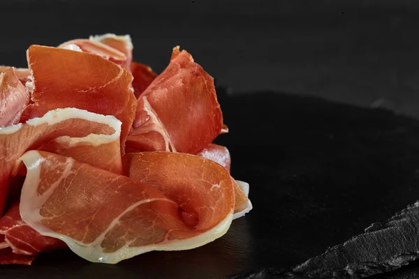 Gurmán, ručně krájený jamon na černé kamenné břidlicové desce na tmavě šedém pozadí. Záběr zblízka. Boční pohled. Kopírovat prostor. — Stock fotografie