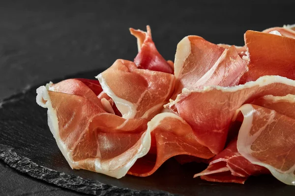 Gurmán, ručně krájený jamon na černé kamenné břidlicové desce na tmavě šedém pozadí. Záběr zblízka. Boční pohled. Kopírovat prostor. — Stock fotografie